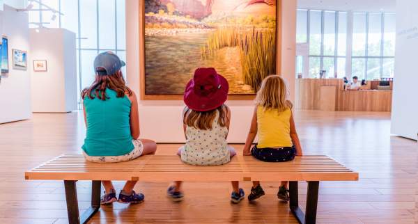 Three kids sitting on a wood bench in front of a landscape painting at the Southern Utah Museum of Art.